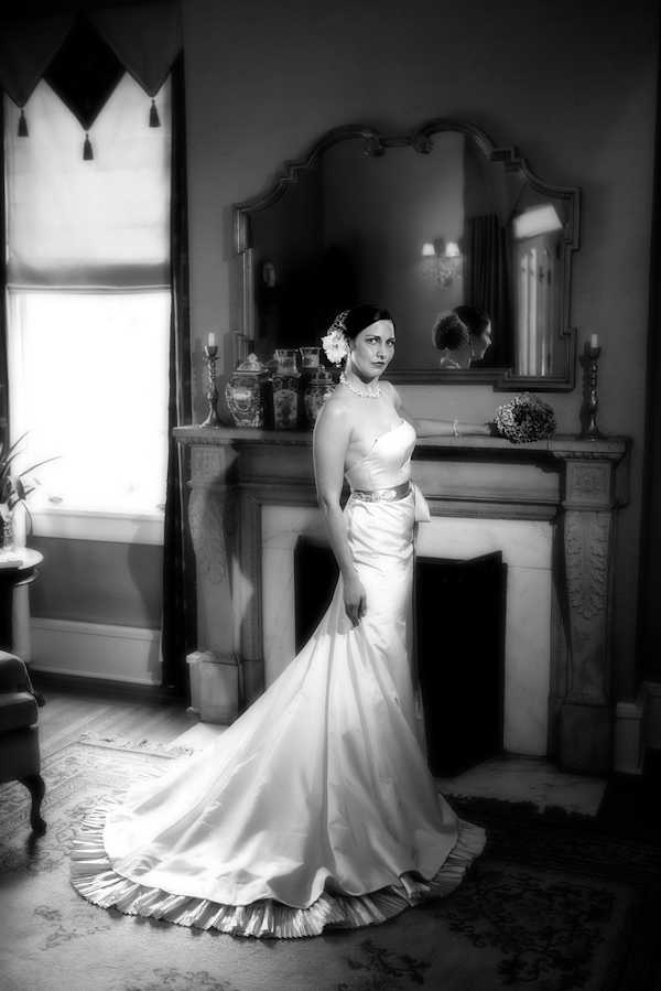 black and white photo - beautiful photo of bride wearing a vintage inspired sheath dress with sash and floral hairpiece standing in front of fireplace - photo by North Carolina based wedding photographers Cunningham Photo Artists